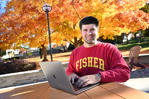A student wearing a Fisher sweatshirt studies at a computer surrounded by fall foliage.