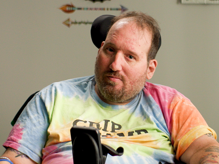 A man in a tie-dyed t shirt, sitting in a power wheel chair.