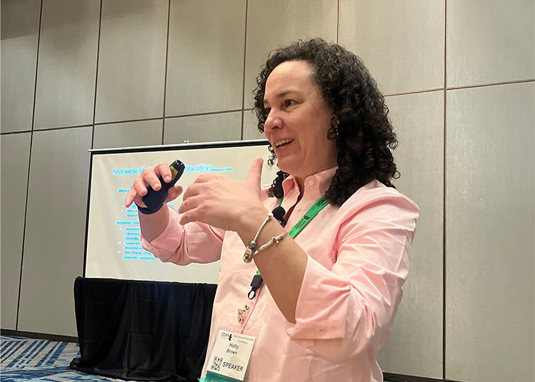 A woman in a light-colored shirt speaks at a conference.