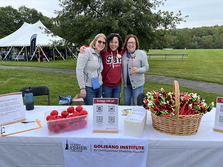 Three women stand behind a table with information about the Golisano Institute for Developmental Disability Nursing. On the table there are baskets filled with pinwheels and stress balls.