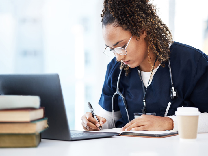 A nurse taking notes at a computer. She has a coffee and a pile of books. She is wearing dark blue scrubs and a stethscope.