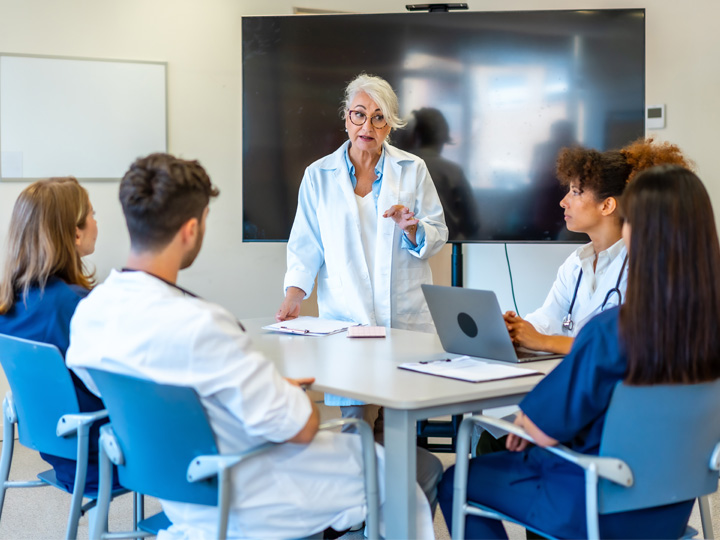 A small group of nurses sits around a table. Another nurse is standing and talking to them.
