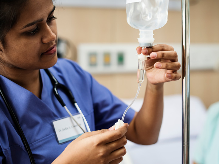A nurse in blue scrubs adjusts and IV in a hospital room.