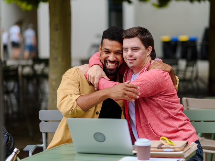 Two men sit at a table in front of an open laptop computer. One is very happy and hugging the other.