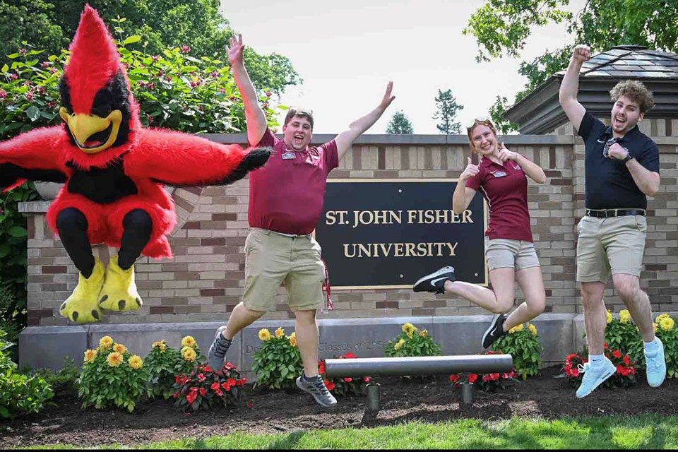 Kurt Schultz (far right) jumps with a group of friends and Cardinal in front of the new St. John Fisher University sign at the campus entrance.
