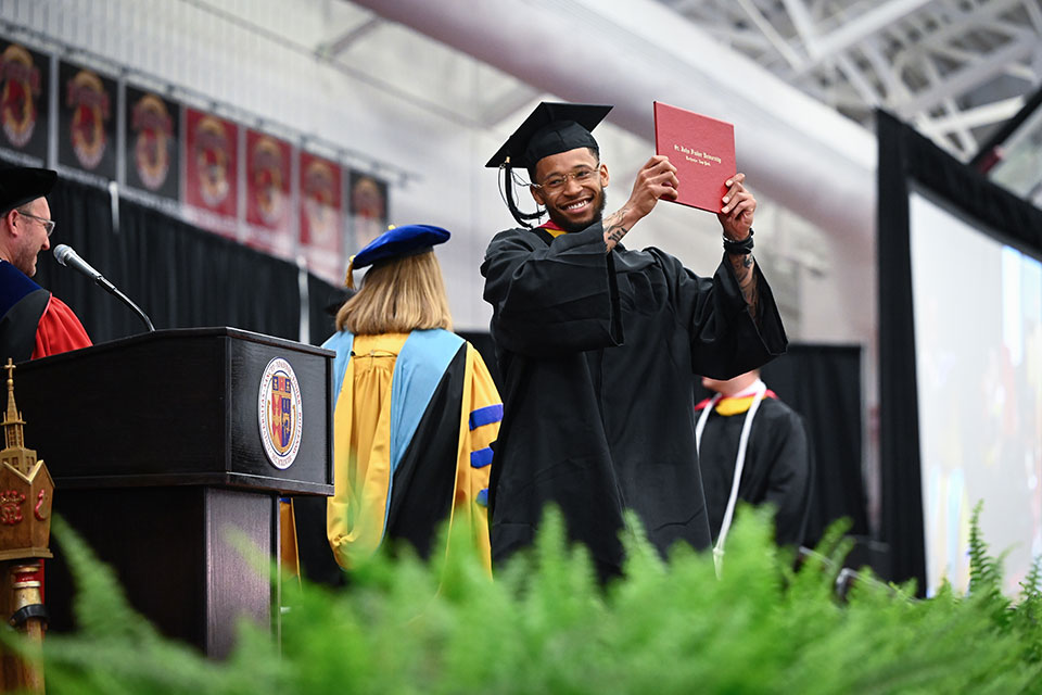 A graduate of the School of Business holds up a diploma while crossing the stage.