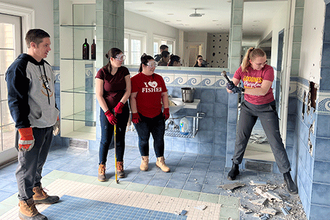 A team of volunteers watch a student knock into a wall with a sledge hammer.