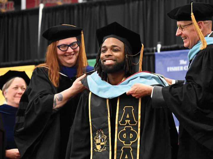 Dean Joellen Maples and a faculty member place an academic hood on a person wearing academic regalia