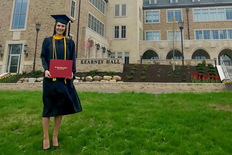 Jessica Bolak poses in front of Kearney Hall in cap and gown holding her degree.