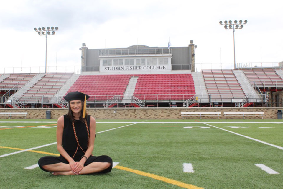 Jessica Colmerauer (Pharm.D.) sits on the turf of Growney Stadium wearing her graduation cap.