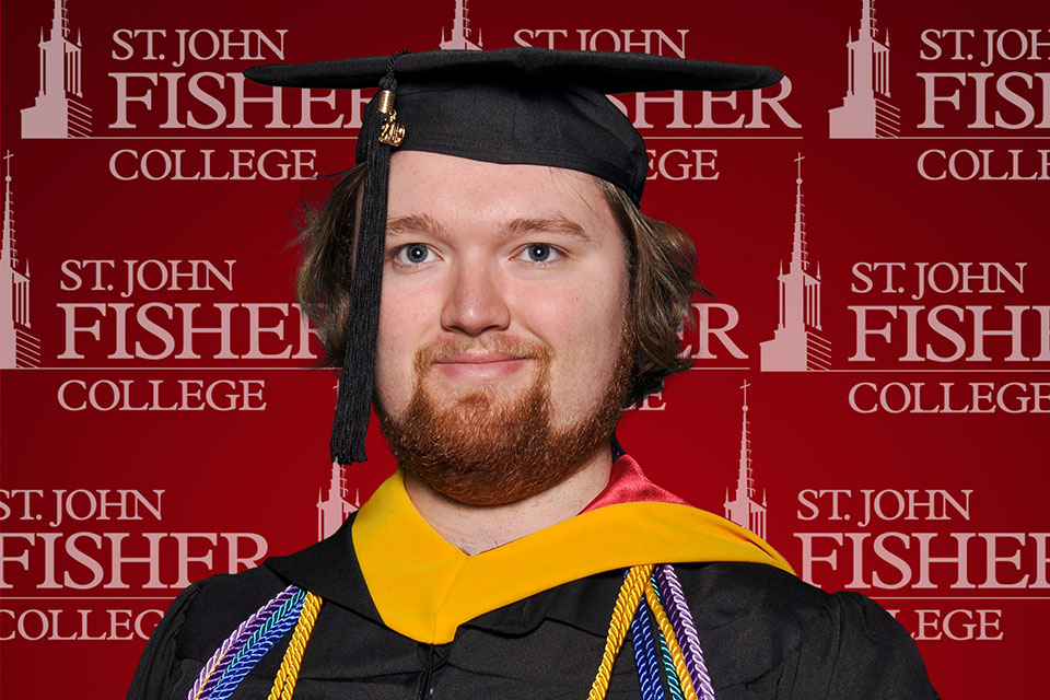 Robert Markwick in cap and gown in front of a St. John Fisher College step and repeat backdrop.