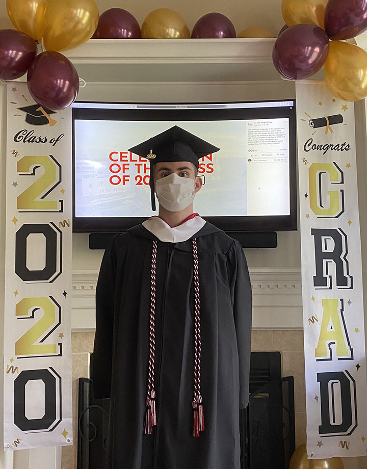 Dylan MacQuoid in cap and gown and mask in front of congratulatory banners.