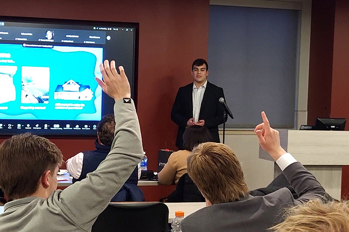 Student raise their hands while a guest lecturer stands at the front of a classroom.