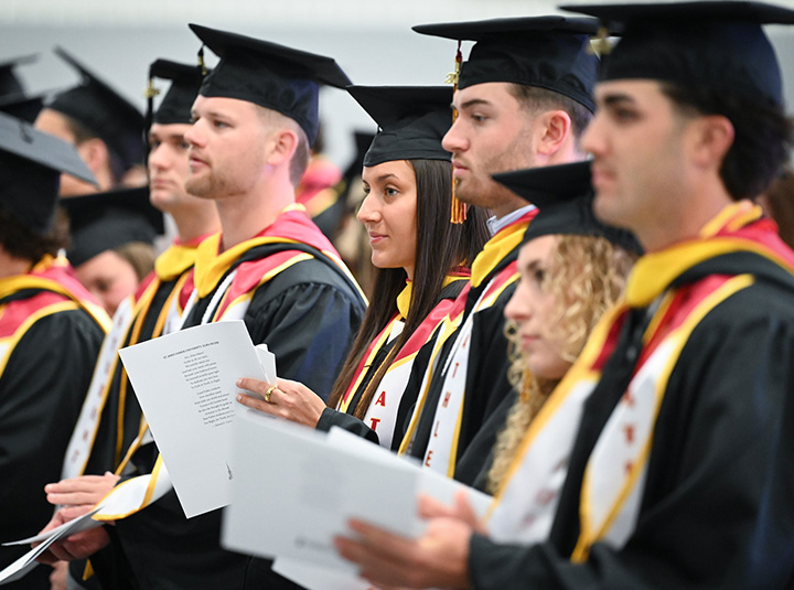 Students in commencement regalia stand with ceremony programs during the Athletics Commencement Ceremony.