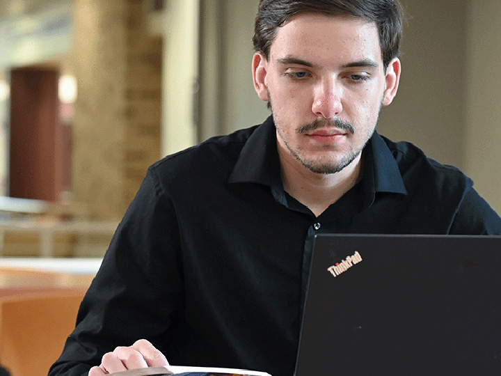 A student studies at a laptop with an open book.