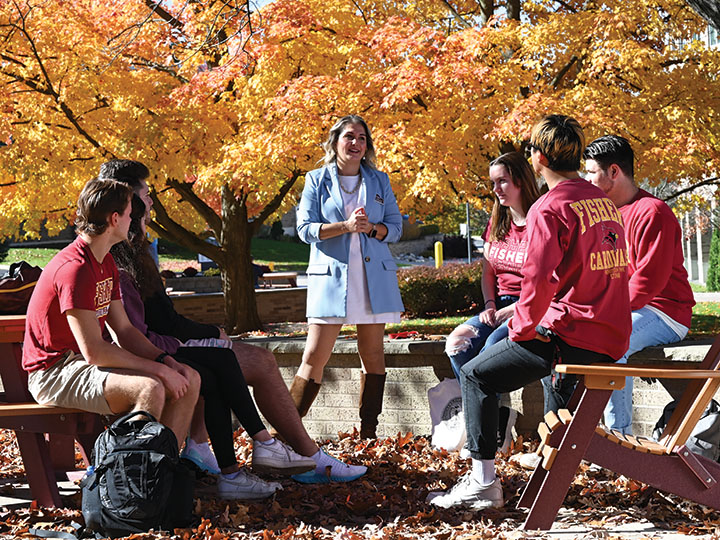 Students sit outside while a professor instructs against a backdrop of fall foliage.