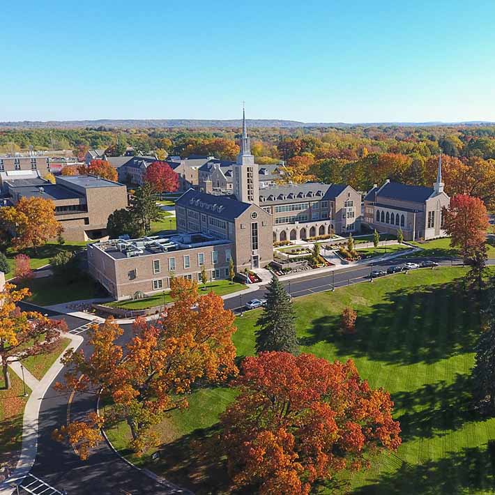 Aerial view of St. John Fisher University with colorful leaves on trees, bright green grassy hills, and sun glistening on buildings.