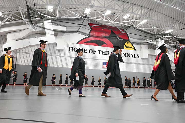 Students process for graduation ceremony in commencement regalia beneath a Cardinal painted on the wall.