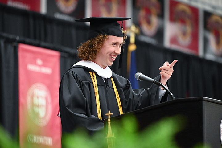 A student in commencement regalia speaks from a podium pointing at graduates.