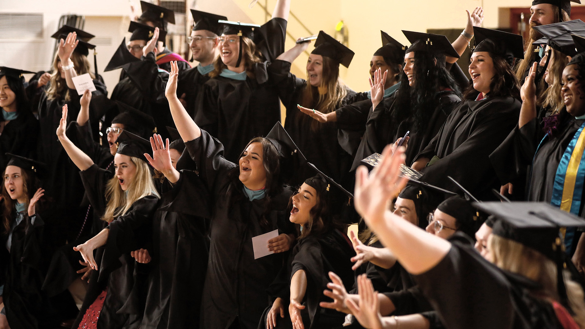 A large group of students wave their hands in Commencement regalia.