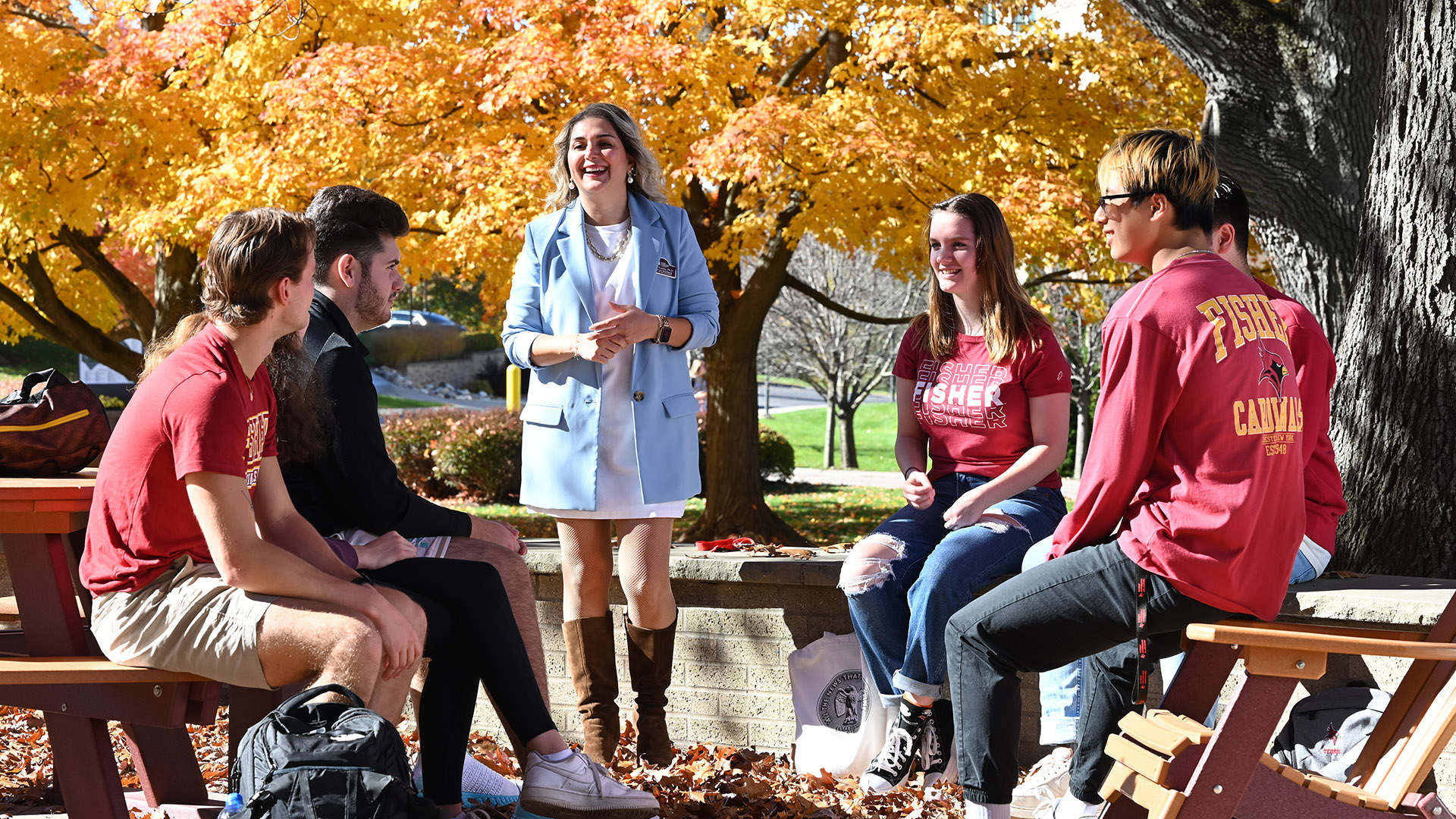 A faculty member talking with students outside on picnic tables in a fall setting.