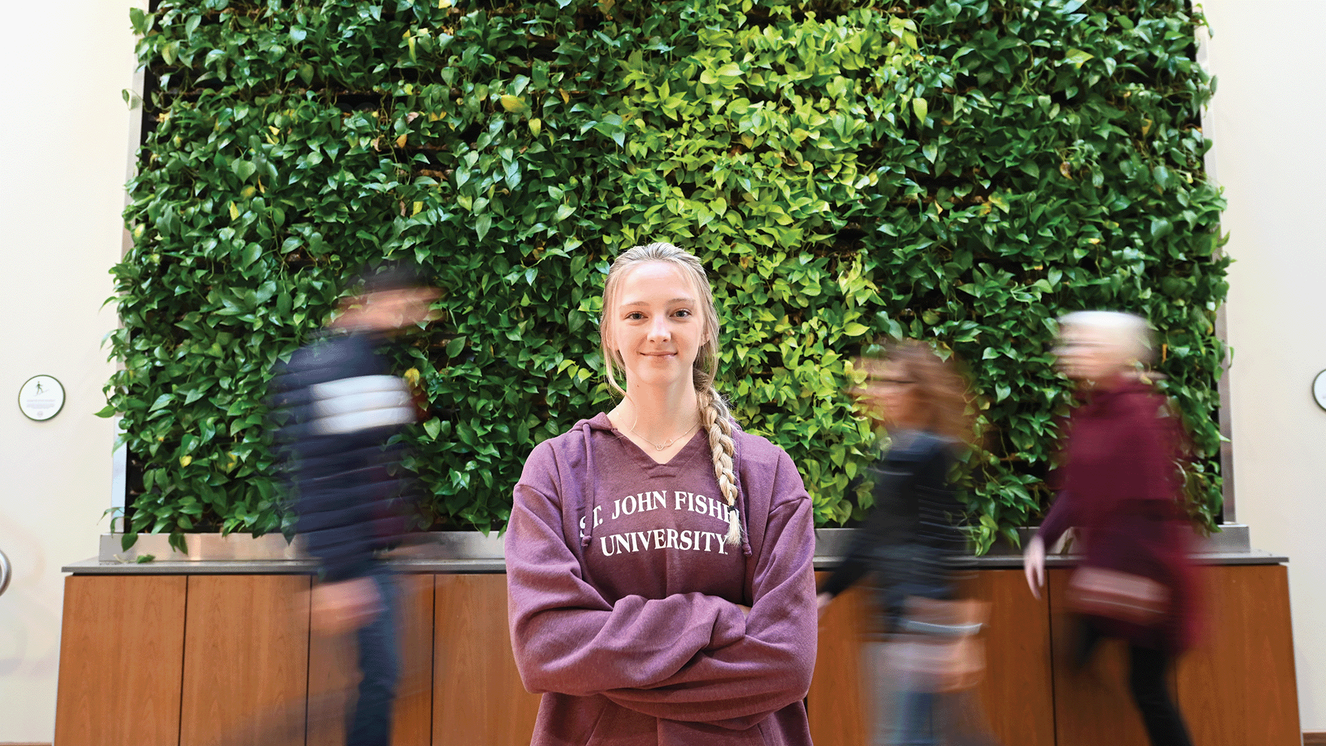 A student stands confidently with arms crossed while students pass by in the background, blurred to show motion.