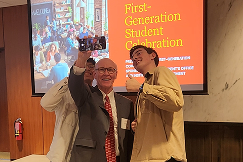 Dr. Gerard Rooney poses for a selfie with two students in front of a screen celebrating First Generation College Student Week.