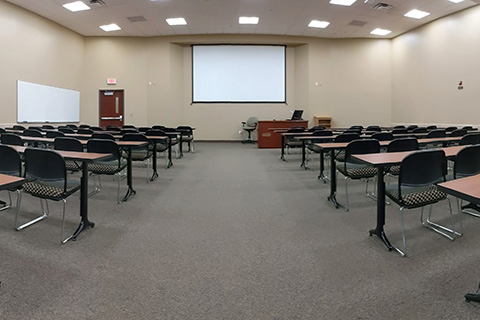 An empty classroom with tables and chairs facing a screen.
