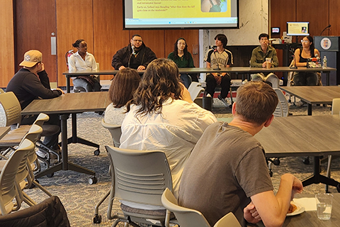 Students on a panel for First-Generation College Student Week speak as other students eat and listen in the audience.