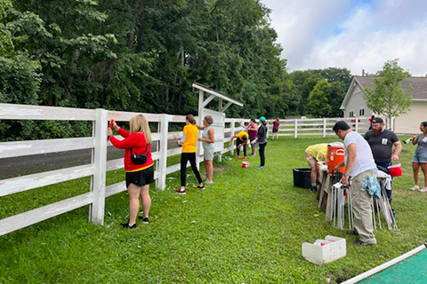 Several people painting and scraping a fence in a field.