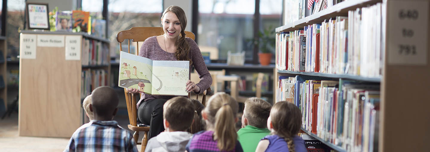 Teacher with book in front of sitting students.