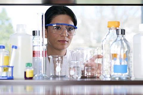 Female pharmacy student stands behind a shelf of bottles and pharmacy equipment.